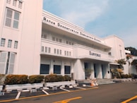 A three-story white building with an art deco architectural style, featuring horizontal lines and large windows. The facade displays the name 'Bappenas' and 'Badan Perencanaan Pembangunan Nasional'. Well-maintained shrubs and potted plants line the building exterior. An empty parking lot with marked spaces is in the foreground, bordered by yellow lines. A clear blue sky is visible above.