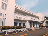 A three-story white building with an art deco architectural style, featuring horizontal lines and large windows. The facade displays the name 'Bappenas' and 'Badan Perencanaan Pembangunan Nasional'. Well-maintained shrubs and potted plants line the building exterior. An empty parking lot with marked spaces is in the foreground, bordered by yellow lines. A clear blue sky is visible above.