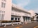 A three-story white building with an art deco architectural style, featuring horizontal lines and large windows. The facade displays the name 'Bappenas' and 'Badan Perencanaan Pembangunan Nasional'. Well-maintained shrubs and potted plants line the building exterior. An empty parking lot with marked spaces is in the foreground, bordered by yellow lines. A clear blue sky is visible above.