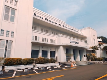 A three-story white building with an art deco architectural style, featuring horizontal lines and large windows. The facade displays the name 'Bappenas' and 'Badan Perencanaan Pembangunan Nasional'. Well-maintained shrubs and potted plants line the building exterior. An empty parking lot with marked spaces is in the foreground, bordered by yellow lines. A clear blue sky is visible above.