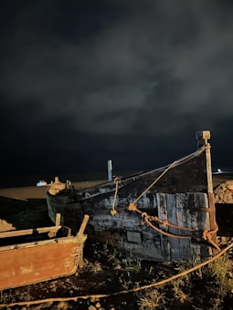 An image depicts an old, weathered fishing boat grounded on a shore during nighttime. The boat is constructed from wood and shows signs of decay, with peeling paint and rusted metal elements. It is anchored with ropes on a rocky and grassy area beside another smaller boat. Dark clouds fill the sky, contributing to a somber and mysterious atmosphere.