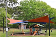 A playground area is surrounded by lush green trees. The play structure is equipped with slides and climbing features, designed for children. Above the playground, colorful orange and blue shade sails provide cover. Nearby, a brick building with a sign indicating it's part of a local council can be seen. People are visible, including children playing and adults nearby.