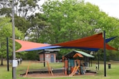 A playground area is surrounded by lush green trees. The play structure is equipped with slides and climbing features, designed for children. Above the playground, colorful orange and blue shade sails provide cover. Nearby, a brick building with a sign indicating it's part of a local council can be seen. People are visible, including children playing and adults nearby.