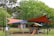 A playground area is surrounded by lush green trees. The play structure is equipped with slides and climbing features, designed for children. Above the playground, colorful orange and blue shade sails provide cover. Nearby, a brick building with a sign indicating it's part of a local council can be seen. People are visible, including children playing and adults nearby.