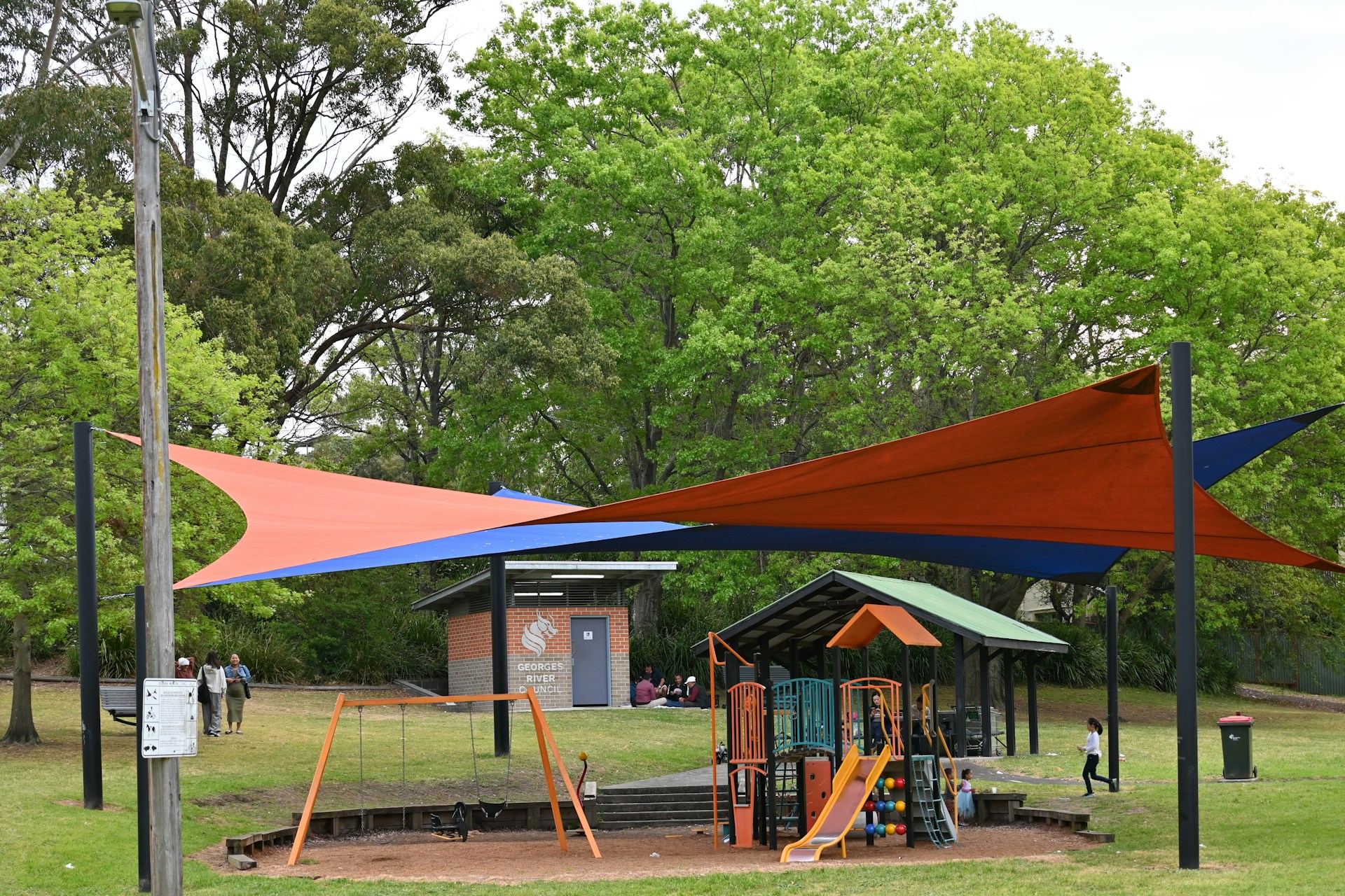 A playground area is surrounded by lush green trees. The play structure is equipped with slides and climbing features, designed for children. Above the playground, colorful orange and blue shade sails provide cover. Nearby, a brick building with a sign indicating it's part of a local council can be seen. People are visible, including children playing and adults nearby.