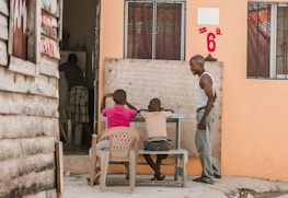 Two children are seated outside on wooden benches at a small desk, engaged with a blackboard that has writing on it. An adult stands nearby, seemingly instructing or observing them. The setting has rustic wooden and painted walls, and the environment appears casual yet educational.