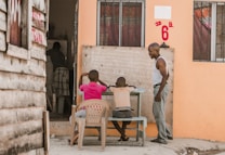 Two children are seated outside on wooden benches at a small desk, engaged with a blackboard that has writing on it. An adult stands nearby, seemingly instructing or observing them. The setting has rustic wooden and painted walls, and the environment appears casual yet educational.