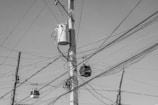 A complex web of electrical wires is strung across several utility poles. A large transformer is mounted on one of the poles, and numerous wires crisscross the scene. The composition is in black and white, highlighting the chaotic arrangement of the cables against a clear sky.