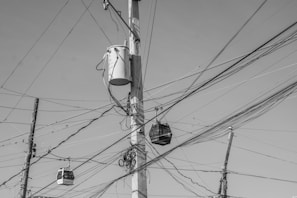 A complex web of electrical wires is strung across several utility poles. A large transformer is mounted on one of the poles, and numerous wires crisscross the scene. The composition is in black and white, highlighting the chaotic arrangement of the cables against a clear sky.