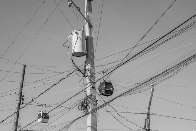 A complex web of electrical wires is strung across several utility poles. A large transformer is mounted on one of the poles, and numerous wires crisscross the scene. The composition is in black and white, highlighting the chaotic arrangement of the cables against a clear sky.