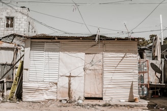 A modest structure constructed with corrugated metal sheets and wood planks, featuring a makeshift door and window. The area is surrounded by electrical wires crisscrossing overhead, with visible wear and some debris on the ground. A small ladder leans against the wall, and in the background, there is an unfinished brick building.