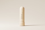 A nasal inhaler is centered against a minimalistic, beige background. The inhaler is cream-colored with sleek, modern typography printed vertically along its body. The lighting accentuates its smooth, cylindrical shape and highlights the subtle shadows around its base.