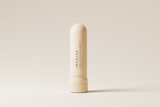 A nasal inhaler is centered against a minimalistic, beige background. The inhaler is cream-colored with sleek, modern typography printed vertically along its body. The lighting accentuates its smooth, cylindrical shape and highlights the subtle shadows around its base.