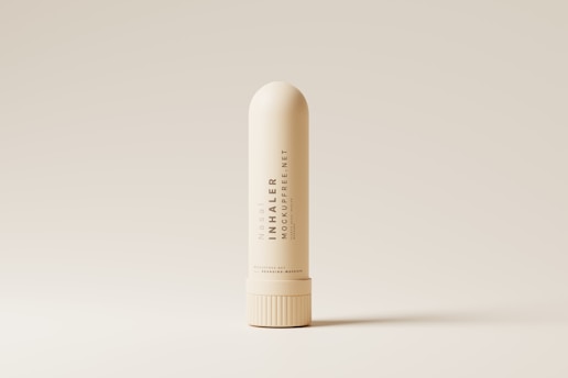 A nasal inhaler is centered against a minimalistic, beige background. The inhaler is cream-colored with sleek, modern typography printed vertically along its body. The lighting accentuates its smooth, cylindrical shape and highlights the subtle shadows around its base.
