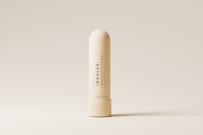 A nasal inhaler is centered against a minimalistic, beige background. The inhaler is cream-colored with sleek, modern typography printed vertically along its body. The lighting accentuates its smooth, cylindrical shape and highlights the subtle shadows around its base.