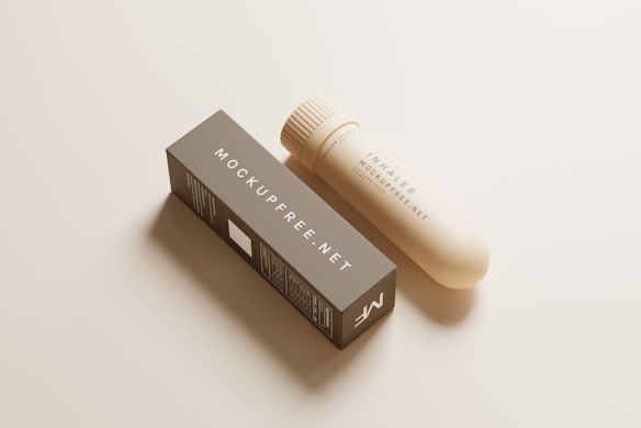 A beige inhaler placed beside a rectangular box labeled with 'MOCKUPFREE.NET'. The inhaler has a cylindrical shape with textured grip near the top. Both items rest on a smooth, light-colored surface.