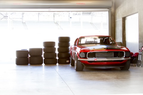 A bright garage scene features a classic red car parked alongside stacks of racing tires. The interior presents a bare concrete floor and walls, with ample lighting coming through a large garage door.