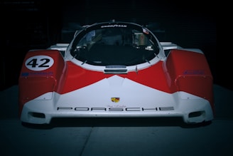 A vintage Porsche race car with a prominent red and white color scheme, featuring the number 42 on its side and various sponsor decals. The car has a sleek, aerodynamic design with a low front end and a well-defined windshield and cockpit.