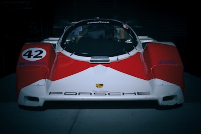 A vintage Porsche race car with a prominent red and white color scheme, featuring the number 42 on its side and various sponsor decals. The car has a sleek, aerodynamic design with a low front end and a well-defined windshield and cockpit.