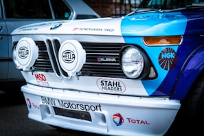 The front of a vintage BMW racing car featuring prominent headlights with 'Hella' branding. The car is adorned with various racing decals and logos, including 'Yokohama Tires', 'STAHL HEADERS', 'BMW Motorsport', and 'Total'. The hood displays a combination of blue and white colors, with the BMW emblem visible on top.
