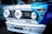 The front of a vintage BMW racing car featuring prominent headlights with 'Hella' branding. The car is adorned with various racing decals and logos, including 'Yokohama Tires', 'STAHL HEADERS', 'BMW Motorsport', and 'Total'. The hood displays a combination of blue and white colors, with the BMW emblem visible on top.