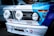 The front of a vintage BMW racing car featuring prominent headlights with 'Hella' branding. The car is adorned with various racing decals and logos, including 'Yokohama Tires', 'STAHL HEADERS', 'BMW Motorsport', and 'Total'. The hood displays a combination of blue and white colors, with the BMW emblem visible on top.