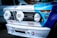 The front of a vintage BMW racing car featuring prominent headlights with 'Hella' branding. The car is adorned with various racing decals and logos, including 'Yokohama Tires', 'STAHL HEADERS', 'BMW Motorsport', and 'Total'. The hood displays a combination of blue and white colors, with the BMW emblem visible on top.