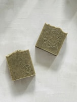 Two rectangular soap bars with an earthy texture are placed on a light backdrop. One soap has a circular emblem with symbols arranged in a circular pattern, while the other displays a simpler circular design. The edges of the soaps are slightly rough, suggesting a handmade quality.