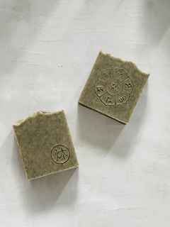 Two rectangular soap bars with an earthy texture are placed on a light backdrop. One soap has a circular emblem with symbols arranged in a circular pattern, while the other displays a simpler circular design. The edges of the soaps are slightly rough, suggesting a handmade quality.
