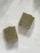 Two rectangular soap bars with an earthy texture are placed on a light backdrop. One soap has a circular emblem with symbols arranged in a circular pattern, while the other displays a simpler circular design. The edges of the soaps are slightly rough, suggesting a handmade quality.