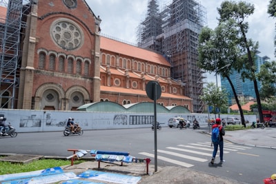 A large, historic church with red brick walls and ornate circular windows is undergoing renovation with scaffolding visible around the towers. Several motorcycles and cars are in motion on the surrounding street, and a few pedestrians are present, one of whom is crossing a crosswalk. Lush green trees frame the scene, and modern buildings are visible in the distance.