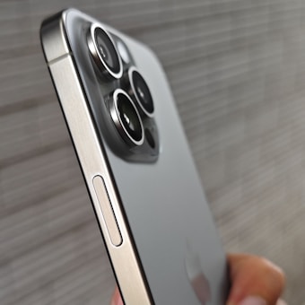 A close-up of a smartphone displaying its sleek metallic edge and prominent camera lenses. The phone's design features multiple camera lenses arranged in a square cluster on its back. The background is blurred, focusing attention on the phone's detail and finish.