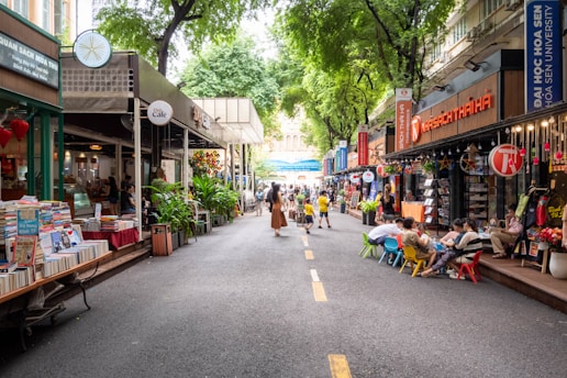A vibrant street scene features an outdoor marketplace lined with bookshops and cafes. People browse books and enjoy leisurely activities, while children are seated on small, colorful chairs. Tall green trees provide shade, creating a lively and inviting atmosphere.
