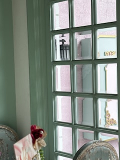A decorative interior space featuring a window with glass panes. The window frame is painted in a soft green color, matching the walls. Through the window, the exterior shows a pale pink textured wall with some architectural details in green and gold. Inside, a vintage-style chair with an ornate floral pattern is partially visible, along with a bouquet of artificial flowers placed in front of it.