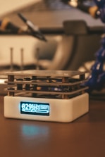 A small electronic device with a digital display sits on a tabletop. The display shows numerical data and the word 'SHUT'. The device has a cubic form with layers of metal, possibly part of a cooling system.