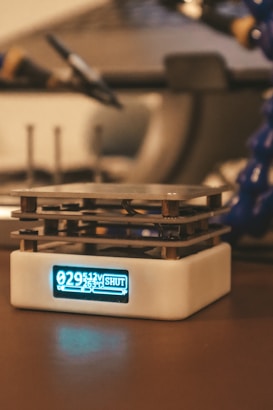 A small electronic device with a digital display sits on a tabletop. The display shows numerical data and the word 'SHUT'. The device has a cubic form with layers of metal, possibly part of a cooling system.