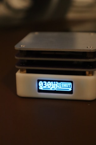 A compact electronic device with a digital display showing the numbers 030, 5.12V, and the word SHUT. It has a white casing with a black screen, and it's placed on a dark surface.