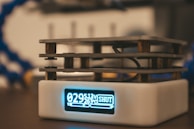 A close-up view of a small electronic device with a digital screen displaying numbers and the word 'SHUT.' The device has a rectangular shape with a metal framework on top and appears to be placed on a flat surface.