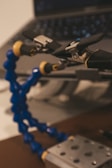 A close-up of a flexible tool with blue and yellow segmented arms, equipped with black and silver clamps, set against a blurred background of a laptop. The focus is primarily on the tool, suggesting it may be used for holding or manipulating small objects.
