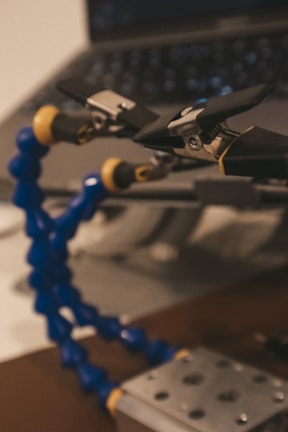 A close-up of a flexible tool with blue and yellow segmented arms, equipped with black and silver clamps, set against a blurred background of a laptop. The focus is primarily on the tool, suggesting it may be used for holding or manipulating small objects.