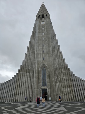 A tall, striking concrete church building with a unique stepped facade and a clock near the top. The entrance features a large, narrow arched window, and people are visible walking in front of the church on a patterned pavement under an overcast sky.