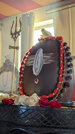 A black Shivling adorned with a white horizontal mark (probably ash), a red dot underneath, and a garland consisting of red and brown beads. There is a golden trishul (trident) placed next to it. Fresh roses and a white flower are placed at the base. The background includes a window with sunlight streaming through, giving a serene and divine ambiance.