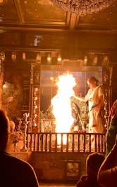 A person dressed in traditional attire performs a ritual involving a large flame in a richly decorated area. The setting includes intricate metalwork and religious icons, with another person observing the ceremony.