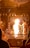 A person dressed in traditional attire performs a ritual involving a large flame in a richly decorated area. The setting includes intricate metalwork and religious icons, with another person observing the ceremony.