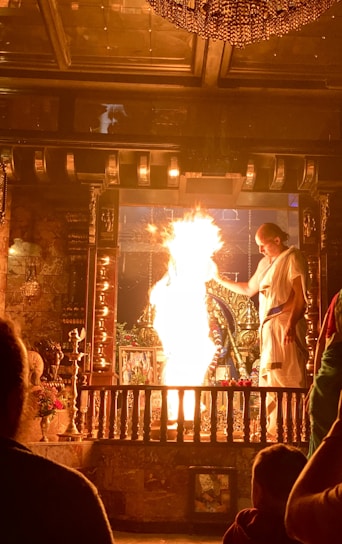 A person dressed in traditional attire performs a ritual involving a large flame in a richly decorated area. The setting includes intricate metalwork and religious icons, with another person observing the ceremony.