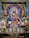 Intricately decorated deity statue adorned with vibrant garlands and flowers, surrounded by framed images of spiritual figures, and flanked by traditional oil lamps. The setting is richly colorful with blue, gold, and floral accents.
