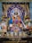 Intricately decorated deity statue adorned with vibrant garlands and flowers, surrounded by framed images of spiritual figures, and flanked by traditional oil lamps. The setting is richly colorful with blue, gold, and floral accents.