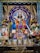 Intricately decorated deity statue adorned with vibrant garlands and flowers, surrounded by framed images of spiritual figures, and flanked by traditional oil lamps. The setting is richly colorful with blue, gold, and floral accents.