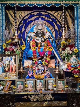 Intricately decorated deity statue adorned with vibrant garlands and flowers, surrounded by framed images of spiritual figures, and flanked by traditional oil lamps. The setting is richly colorful with blue, gold, and floral accents.