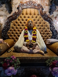 A statue of a seated figure dressed in traditional attire with a garland around the neck, placed on an ornate cushioned seat with a carved wooden backrest. The background features a tiled wall with a marble finish. Flower bouquets are arranged at the base of the seat.